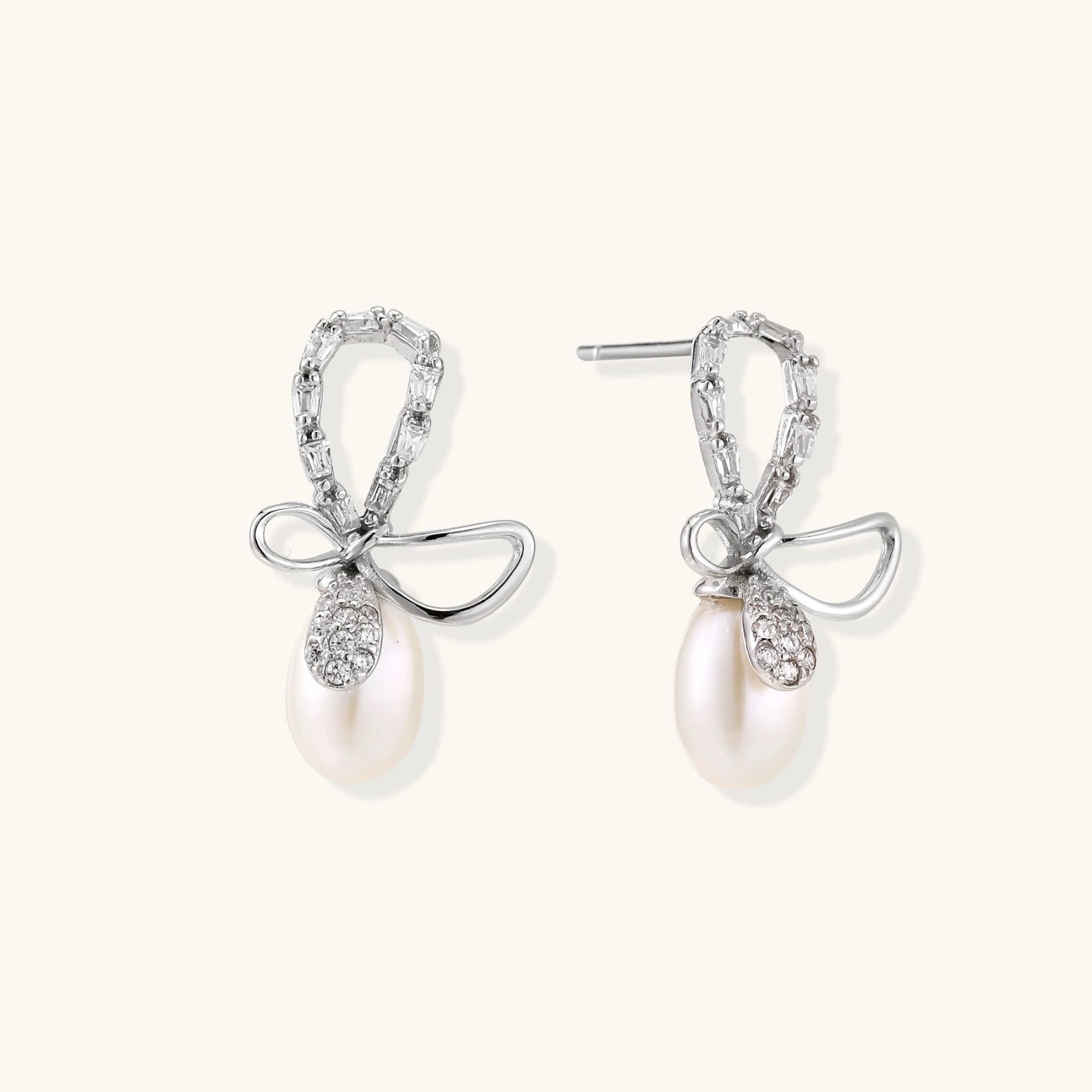 Silver bow-shaped stud earrings with baguette crystals and dangling white freshwater pearls on cream background